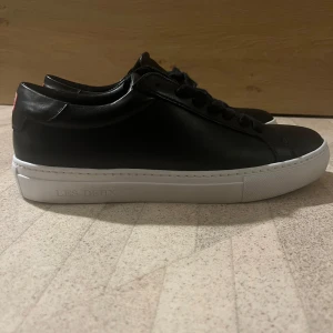  Sneakers från Les Deux - Snygga svarta sneakers från Les Deux med vit sula och klassisk snörning. Perfekta för en stilren look. 