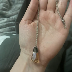 Halsband med droppformad kristall - Elegant halsband med en droppformad, facetterad kristall i ljusbrun nyans. Kristallen är fäst vid en silverkedja och dekorerad med små, glittrande stenar vid infästningen. Perfekt för att ge en touch av glamour till din outfit.