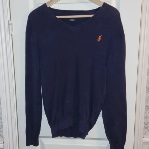 Mörkblå tröja från Ralph Lauren - Säljer en stilren mörkblå tröja från Ralph Lauren i Pima Cotton. Tröjan har en v-ringad design och ett broderat orange logotyp på bröstet. Perfekt för en elegant och avslappnad look. Tyvärr lite solblekt på ena axeln ( se foto), därav priset.