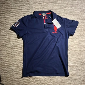 Mörkblå pikétröja från Polo Ralph Lauren - Snygg mörkblå pikétröja från Polo Ralph Lauren med röd broderad logga på bröstet och nummerdetalj på ärmen. Tröjan har en klassisk krage och knappar framtill. Perfekt för en stilren look. Pikén är helt ny som du kan se på lappen. Jag säljer den för att den var lite för liten i storlek