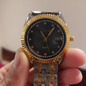 Guld och silverfärgad klocka med mörkgrå urtavla - Elegant klocka med en blå urtavla och datumvisning. Klockan har en guldfärgad boett med glittrande stenar runt urtavlan. Armbandet är i en kombination av guld och silver, vilket ger en lyxig känsla.