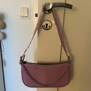 Lila axelväska med kedjedetalj - Snygg lila axelväska med en cool kedjedetalj och justerbar axelrem. Väskan har en dragkedja upptill och ett rymligt innerfack. Perfekt för att ge din outfit en färgklick! Det finns även en liten gullig spegel inuti väskan! Köpt för 200kr!