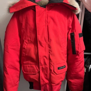  Canada Goose jacka i röd  - Säljer en röd dunjacka från Canada Goose med pälsfodrad huva och dragkedja. Jackan har en patch på ärmen och flera praktiska fickor. Perfekt för kalla vinterdagar.