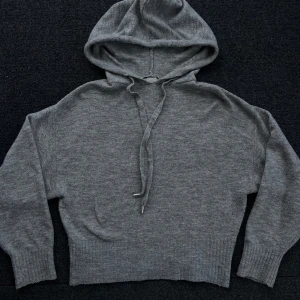 Grå stickad hoodie - Mysig grå stickad hoodie med en smickrande passform och långa ärmar. Perfekt för kyliga dagar med sin bekväma huva och ribbade muddar. En stilren och enkel design som passar till det mesta.