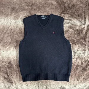 Polo Ralph Lauren - Säljer en stilren mörkblå stickad väst från Polo Ralph Lauren. Västen har en v-ringad design och är tillverkad i 100% bomull. Perfekt för lager-på-lager-stil och har en liten broderad logga på bröstet.