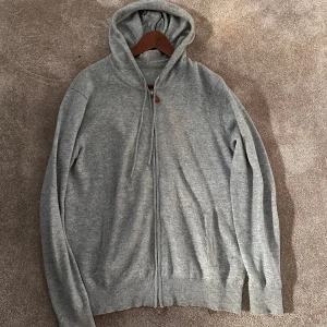 Grå hoodie med dragkedja - Säljer en stilren grå hoodie med dragkedja och huva. Perfekt för kyliga dagar med sin mjuka och bekväma passform. Ribbstickade muddar vid ärmslut och nederkant ger en snygg finish.