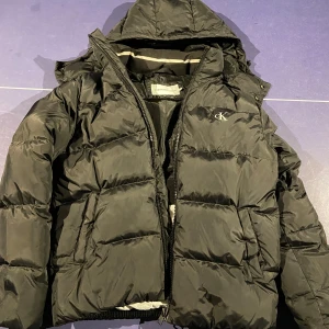 Svart pufferjacka från Calvin Klein - Säljer en svart pufferjacka från Calvin Klein i storlek M med huva och dragkedja. Jackan är perfekt för kyliga dagar och har en stilren design med CK-logga på bröstet. Inga skador förutom ett litet hål längst ner på ena armen som går att sy ihop