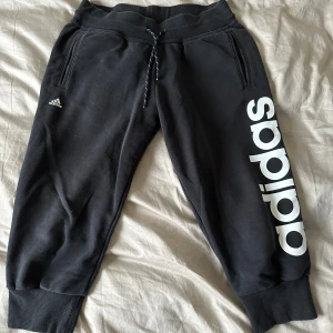Capri mjukisbyxor från Adidas - Säljer ett par svarta capri mjukisbyxor från Adidas med vit logga längs benet. Byxorna har resår i midjan och snörning för justerbar passform. Tillverkade i en mjuk bomulls- och polyesterblandning, perfekta för en avslappnad stil.