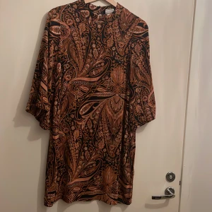 Mönstrad klänning från H&M x Richard Allan - Snygg klänning med ett unikt mönster i svart och orange från H&M x Richard Allan. Klänningen har trekvartsärmar och en lös passform, perfekt för en stilfull look. Materialet känns mjukt och bekvämt mot huden.