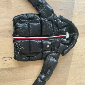 Svart dunjacka från Moncler - Snygg svart dunjacka från Moncler med en iögonfallande röd och vit dragkedja. Jackan har en quiltad design och en praktisk huva. Perfekt för kyliga dagar! Det är ett litet slitage på en av ränderna. 