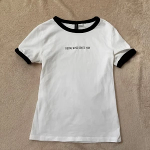Vit t-shirt med svart text - Snygg vit t-shirt med svart text 'BEING KIND SINCE 1989' och svarta kanter vid hals och ärmar. Använd 1 gång, bra skick strl XS.