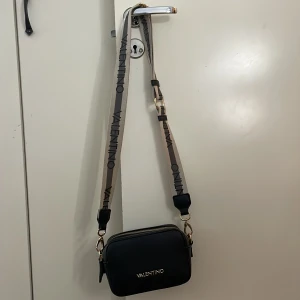 Svart axelväska från Valentino - Snygg svart axelväska från Valentino med justerbar rem och guldfärgade dragkedjor. Väskan har flera fack för praktisk förvaring och en stilren design med logotyp på framsidan. Väskan är i väldigt fint skick och är köpt i början av 2024 för 1300 kr. Säljer för 900 kr då jag snabbt vill bli av me den då den inte används längre. Priset kan diskuteras vid snabb affär, skriv för frågor och funderingar