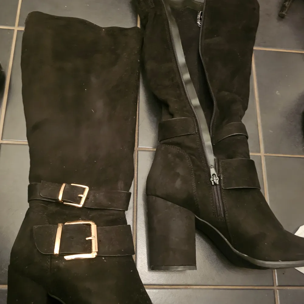 Snygga svarta stövlar i mocka med hög klack och dekorativa spännen. De har en praktisk dragkedja på sidan för enkel på- och avtagning. Perfekta för höst och vinter.. Kengät.