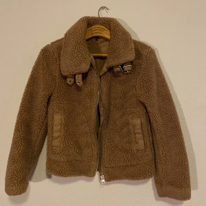 Brun teddyjacka med dragkedja - Mysig brun teddyjacka från Topshop med dragkedja och spännen vid kragen. Perfekt för kyliga dagar med sin fluffiga och varma design. Jackan har långa ärmar och två fickor framtill.