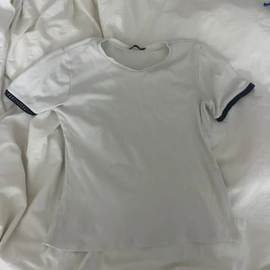 Vit t-shirt från Zara - Säljer en stilren vit t-shirt från Zara med svarta detaljer på ärmsluten. Perfekt för en enkel och klassisk look. Bra skick, inte använd många gånger alls.