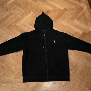 Svart hoodie med dragkedja från Ralph Lauren - Säljer en stilren svart hoodie från Ralph Lauren med dragkedja och huva. Den har en liten broderad logotyp på bröstet och långa ärmar. Perfekt för en avslappnad look. Priset kan diskuteras.