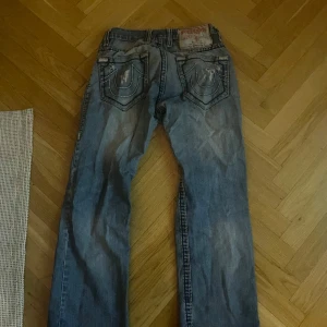 Blå jeans från True Religion - Säljer ett par blå jeans från True Religion med slitna detaljer och broderade bakfickor. Jeansen har en klassisk femficksdesign och knappgylf. Perfekta för en avslappnad stil.