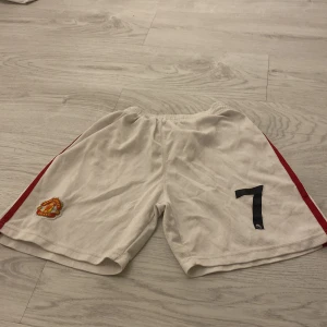 Vita fotbollsshorts med röd detalj - Snygga vita fotbollsshorts med röda ränder längs sidorna och ett tryckt nummer 7 på framsidan. Perfekta för fotbollsträningen eller matchen. Elastisk midja för bekväm passform.