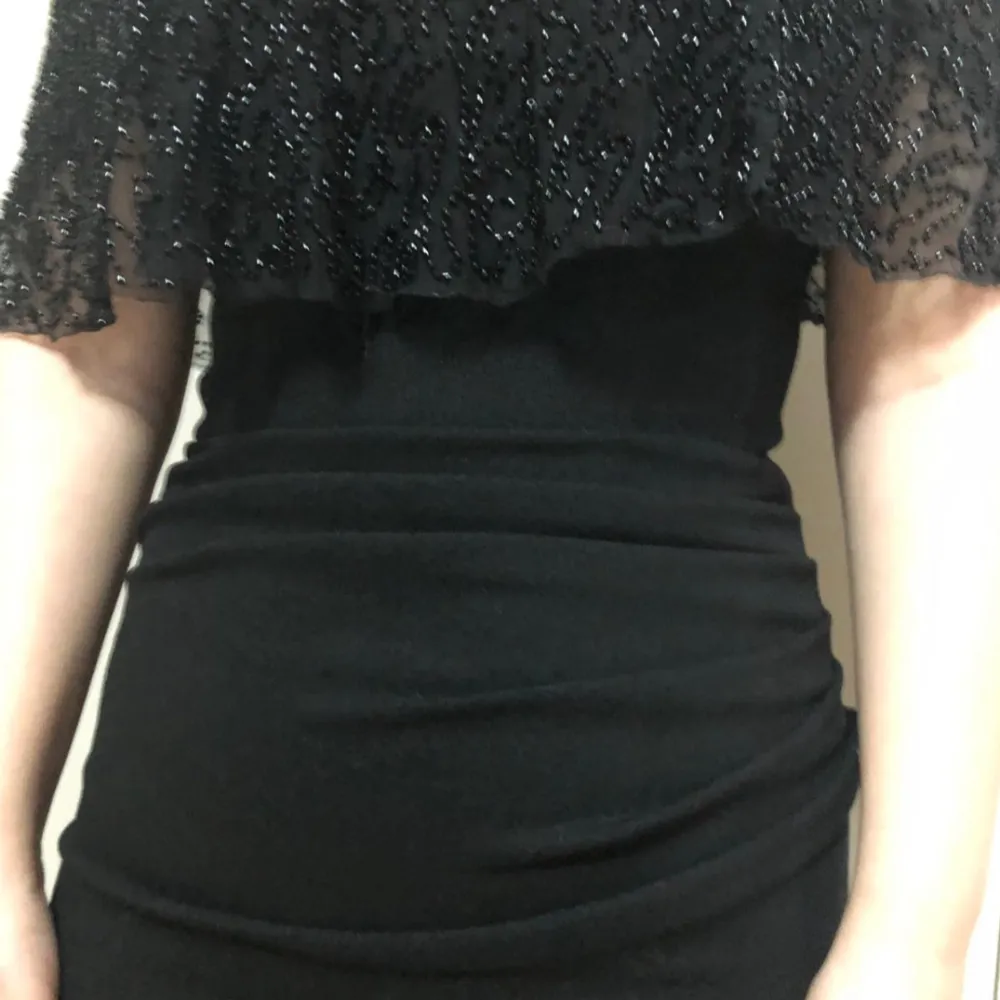 Elegant svart offshoulder klänning med glittriga detaljer och volang. Klänningen har en tight passform och är perfekt för festliga tillfällen.. Mekot.