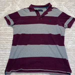 Randig pikétröja från Henri Lloyd - Snygg randig pikétröja från Henri Lloyd i vinrött och grått. Tröjan har korta ärmar och en klassisk krage med knappar. Perfekt för en avslappnad stil.