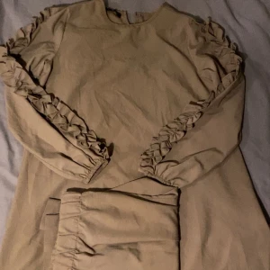 Beige klänning med volangdetaljer - Snygg beige klänning med långa ärmar och volangdetaljer längs ärmarna. Perfekt för en stilren look. Den har en byxa ifall man vill ha den som set och man kan ha den som klänning.