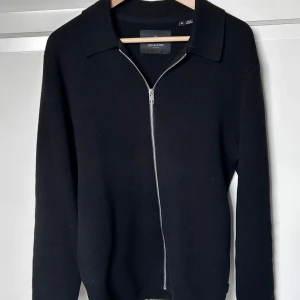 Svart tröja med dragkedja från Jack & Jones - Snygg svart tröja från Jack & Jones med dragkedja framtill. Tröjan har en klassisk krage och långa ärmar, perfekt för en stilren look. Passar bra till både vardag och mer uppklädda tillfällen. Hör av er vid intresse/ frågor!
