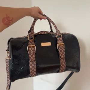 Svart resväska med bruna detaljer - Snygg svart resväska från River Island med bruna handtag och detaljer. Väskan har ett präglat mönster och en praktisk dragkedja. Perfekt för resor och weekendutflykter.