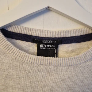 Grå tröja från Smog - Grå tröja från Smog med regular fit. Tröjan har långa ärmar och ribbade muddar vid ärmslut och nederkant. Lite nopprig baksida nederkant.