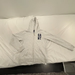 Grå hoodie från GANT - Säljer en stilren grå hoodie från GANT med dragkedja och huva. Tröjan har långa ärmar och en broderad logotyp på bröstet. Perfekt för en avslappnad look. Säljer varan för att den är för liten.