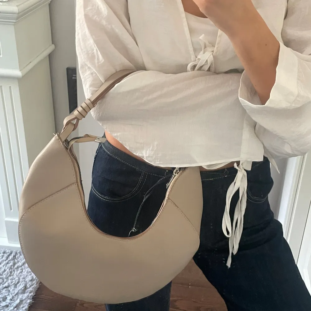 Säljer en superfin beige axelväska i syntetmaterial. Den har en unik halvmåneform och en praktisk dragkedja upptill. Perfekt för både vardag och fest! 👜✨. Väskor.