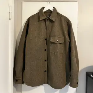 Säljer en snygg brun overshirt från H&M i storlek L. Den har en klassisk krage och knäppning framtill med svarta knappar. En bröstficka med knappdetalj ger en stilren look. Perfekt för höst och vår, och funkar både som jacka och skjorta. Materialet känns robust och håller värmen bra. Passar perfekt för en casual stil!