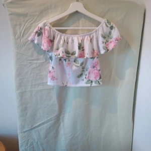 Blommig offshoulder topp - Säljer en superfin offshoulder topp med blommigt mönster. Den är vit med stora rosa blommor och gröna blad. Toppen har en volangdetalj som ger den en romantisk känsla. Perfekt för sommardagar eller en festlig kväll!
