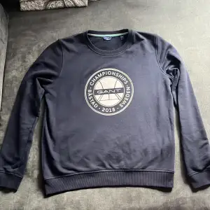 Säljer en snygg svart sweatshirt från GANT med ett coolt tryck på framsidan som säger 'CHAMPIONSHIPS BÅSTAD 2018 SWEDEN'. Tröjan är i storlek M och är gjord av 90% bomull och 10% polyester, vilket gör den både bekväm och hållbar. Perfekt för en avslappnad stil!