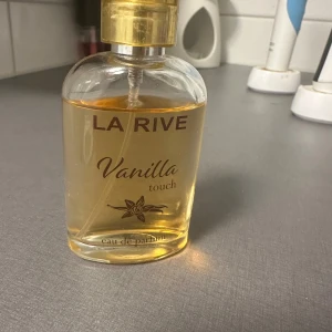 La Rive Vanilla Touch Eau de Parfum - Säljer en flaska La Rive Vanilla Touch Eau de Parfum. Flaskan är genomskinlig med en gyllene kork och innehåller en ljusgul doftvätska. Den har en söt och varm vaniljdoft som är perfekt för dagligt bruk eller speciella tillfällen. Flaskan är elegant och enkel, vilket gör den till ett fint tillskott på sminkbordet.