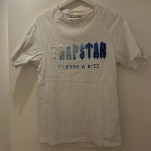 Vit t-shirt från Trapstar - Säljer en vit t-shirt från Trapstar med texten 'TRAPSTAR IT'S A SECRET' i blå och grå nyanser på framsidan. T-shirten är kortärmad och har en klassisk passform. Perfekt för en avslappnad stil.