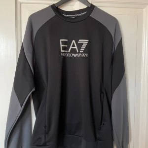 Svart och grå tröja från Emporio Armani - Snygg svart och grå tröja från Emporio Armani med EA7-logga på bröstet. Tröjan har långa ärmar och en bekväm passform, perfekt för en stilren look.