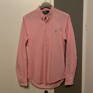 Rosa randig skjorta från Polo Ralph Lauren - Snygg rosa och vit randig skjorta från Polo Ralph Lauren i slim fit. Skjortan har långa ärmar och klassiska knappar framtill. Den ikoniska loggan är broderad på bröstet i blått. Perfekt för en stilren look! Bra skick! Storlek S men passar också M. Mitt pris: 499kr! 