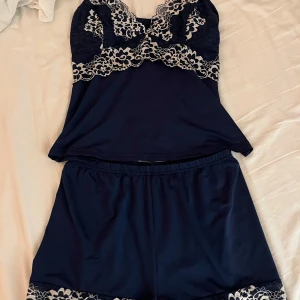 Mörkblå pyjamas med spetsdetaljer - Snygg mörkblå pyjamas med vackra spetsdetaljer i blommönster. Toppen har smala axelband och spets längs bysten, medan shortsen har spetskant vid benen. Perfekt för en bekväm och stilfull natt.
