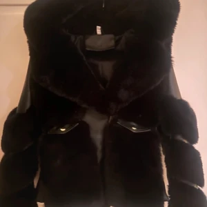 Fluffig Svart pälsjacka  - Snygg svart pälsjacka med läderdetaljer ackan har en lyxig look med en stor krage och är perfekt för kyligare dagar.
