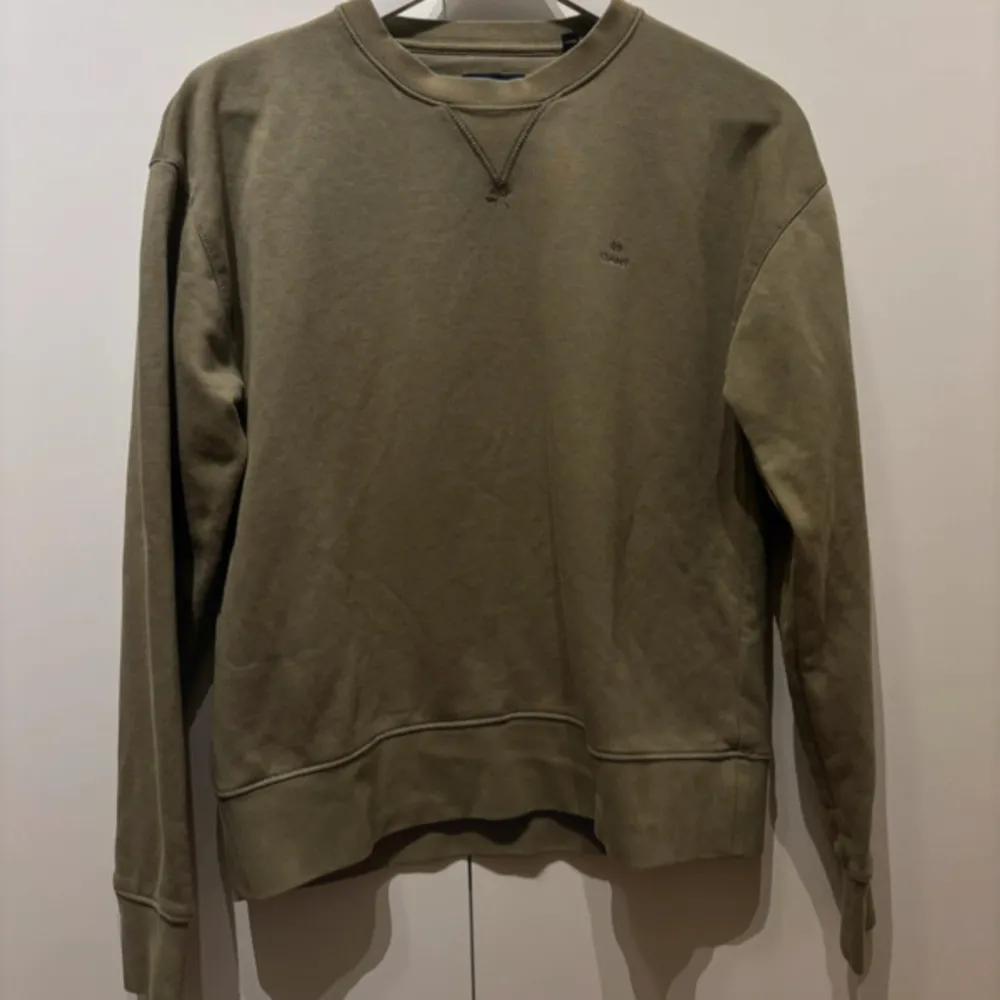 Säljer en stilren olivgrön sweatshirt från GANT. Tröjan har en klassisk rund halsringning och långa ärmar. Perfekt för en avslappnad look. Passar bra till både jeans och chinos.. Neuletakit & Villapaidat.