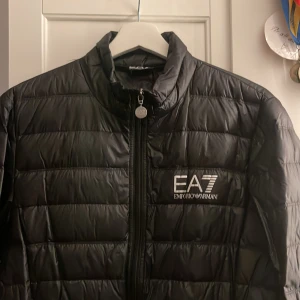 Svart dunjacka från Emporio Armani - Snygg svart dunjacka från Emporio Armani med EA7-logga på bröstet. Jackan har en dragkedja framtill och är quiltad för extra värme. Perfekt för kyligare dagar.