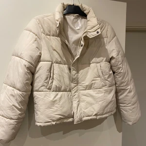 Beige pufferjacka från Gina Tricot - Säljer en beige pufferjacka från Gina Tricot. Jackan har en quiltad design med dragkedja och knappar framtill. Perfekt för kyligare dagar med sin varma och bekväma passform.