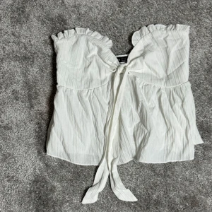 Vit axelbandslös topp med volang - Säljer en söt vit axelbandslös topp med volangdetaljer och knyt framtill. Perfekt för sommardagar och festliga tillfällen. Toppen har en luftig och bekväm passform.