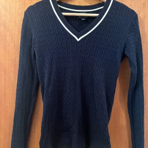 Mörkblå kabelstickad tröja från Tommy Hilfiger - Snygg mörkblå kabelstickad tröja från Tommy Hilfiger i pima bomull. Tröjan har en v-ringad hals med vit kant och långa ärmar. Perfekt för en stilren look.