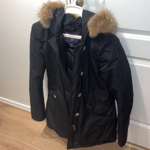 Svart parkajacka från Woolrich - Säljer en svart parkajacka från Woolrich med päls på huvan. Jackan har dragkedja och knappar framtill samt praktiska fickor. Använd ett fåtal gånger under en vinter, resten av tiden har den hängt i förrådet. Köpt för ca 8000 hos Thernlunds och har EJ kvitto.