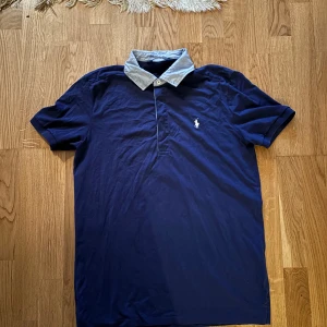 Blå pikétröja från Ralph Lauren - Säljer en stilren blå pikétröja från Ralph Lauren med en kontrasterande ljusblå krage. Tröjan har korta ärmar och en klassisk knappslå framtill. Perfekt för en avslappnad men ändå elegant look.