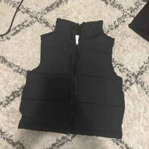 Svart dunväst från Gina Tricot - Säljer en stilren svart dunväst från Gina Tricot. Västen har en praktisk dragkedja framtill och är perfekt för kyligare dagar. Den är enkel att matcha med olika outfits och ger en snygg look.