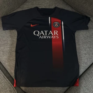 Svart PSG fotbollströja - Snygg svart PSG fotbollströja från Nike med röd och vit detalj på framsidan. Tröjan har korta ärmar och klubbens emblem på bröstet. Perfekt för fans av Paris Saint-Germain!