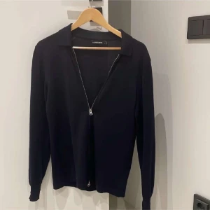 Svart tröja med dragkedja från J.Lindeberg - Snygg svart tröja från J.Lindeberg med dragkedja framtill. Tröjan har långa ärmar och en klassisk krage, perfekt för en stilren look. Passar bra till både vardag och mer uppklädda tillfällen.