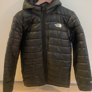 Svart pufferjacka från The North Face - Snygg svart halvtjock  pufferjacka från The North Face med huva och dragkedja framtill. Något liten i storleken enligt mig.